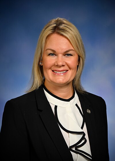 A woman with short blonde hair and wearing a black suit jacket and white blouse poses for a portrait in front of a blue background.